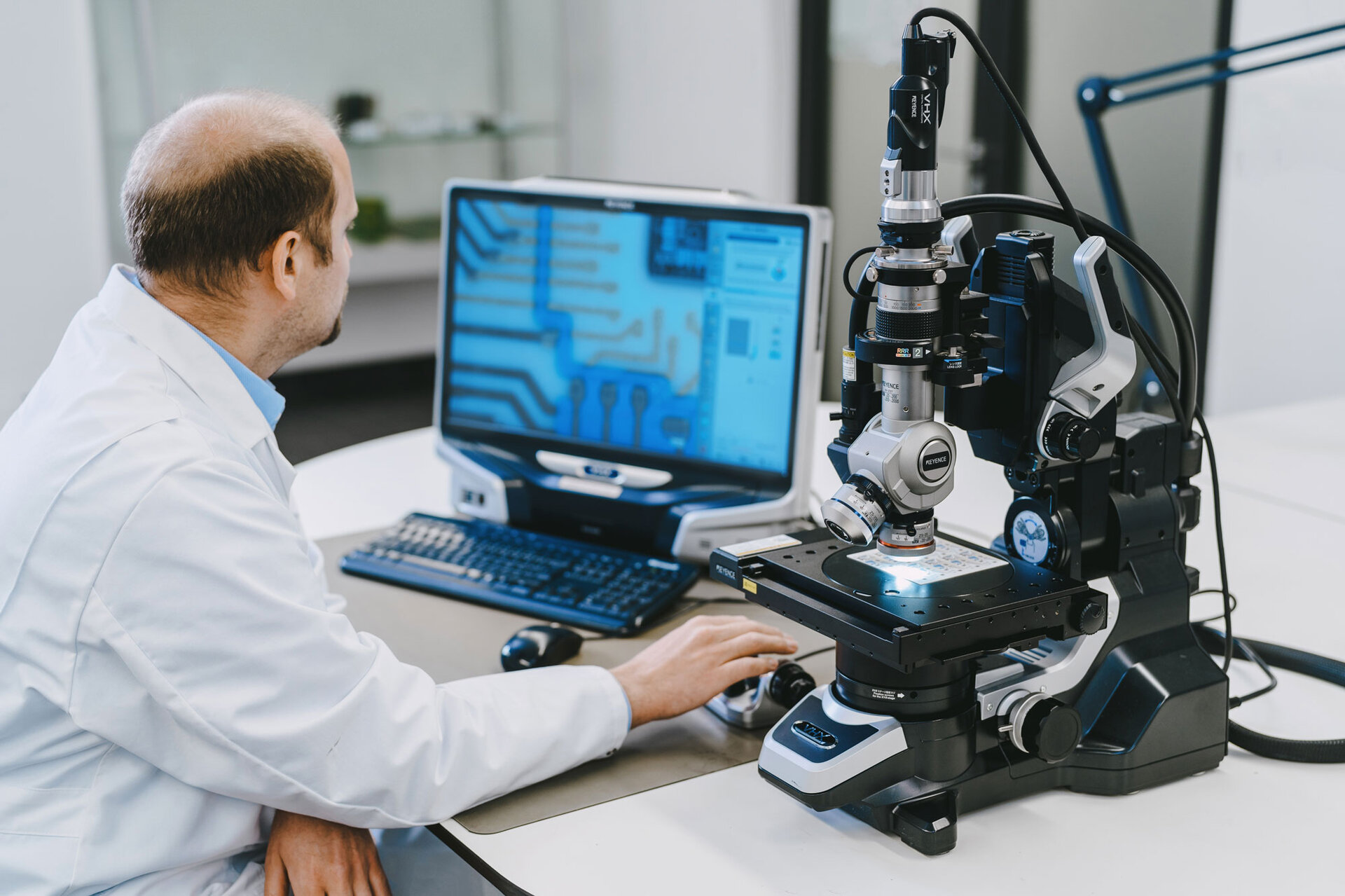 Ein Labormitarbeiter sitzt am Schreibtisch vor einem Mikroskop und führt eine visuelle Schadensanalyse durch, um die Verunreinigung der elektronischen Baugruppe zu bestimmen. | © @The Sour Cherry Fotografie - Michaela Curtis