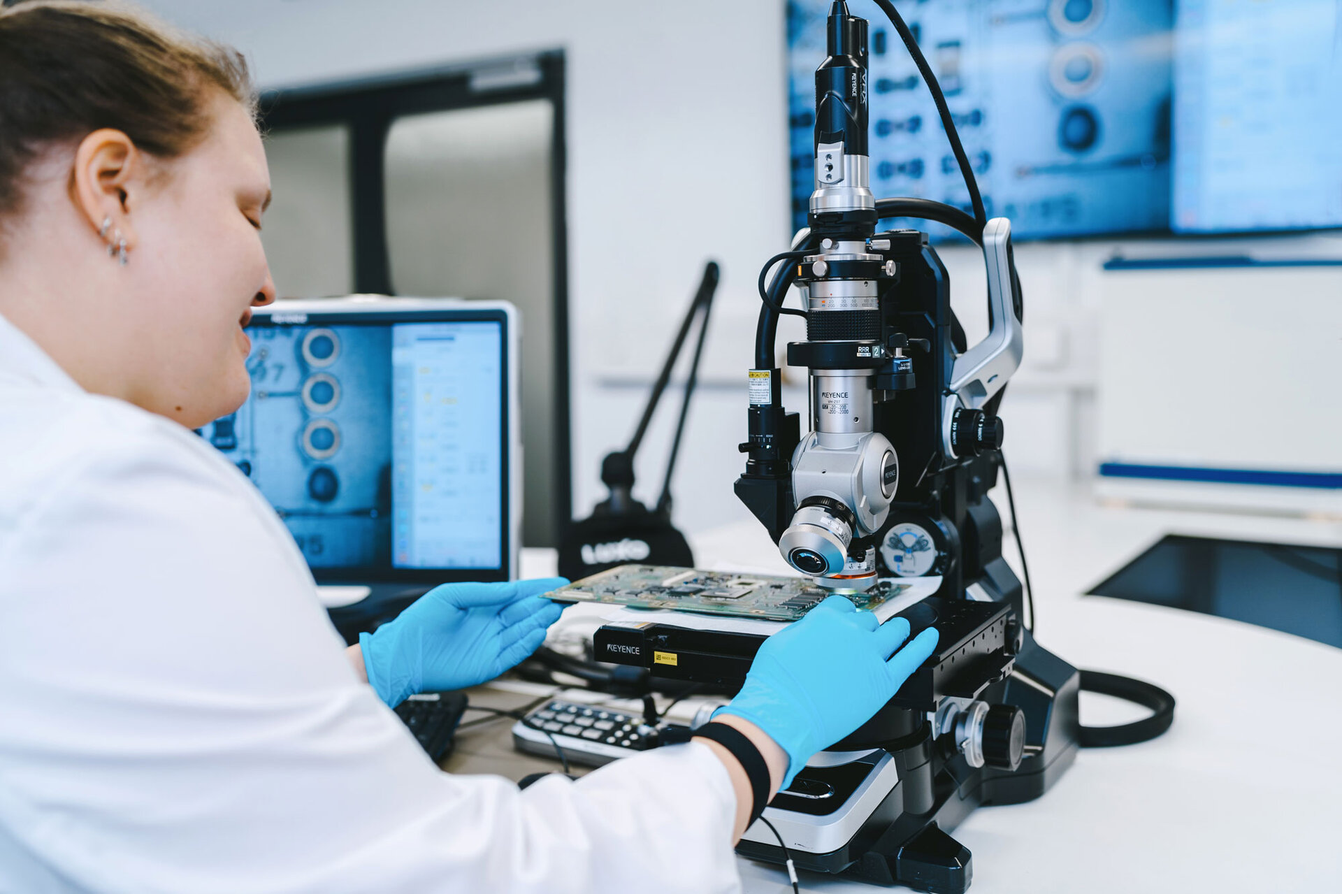 Eine grüne Leiterplatte wird von einer Labormitarbeiterin des Analysezentrums von Zestron durch ein Keyence Mikroskop betrachtet.  | © @The Sour Cherry Fotografie - Michaela Curtis