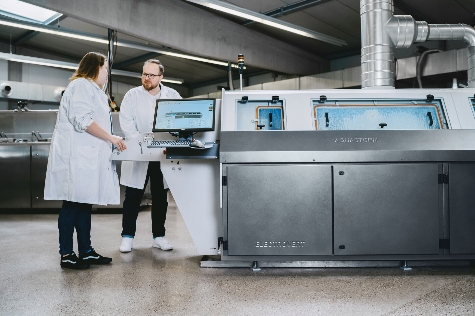 Zwei Ingenieure stehen vor einer Inline-Reinigungsmaschine im Maschinen-Testzentrum und bereiten den Reinigungsprozess für die elektronischen Bauteile vor. | © @The Sour Cherry Fotografie - Michaela Curtis