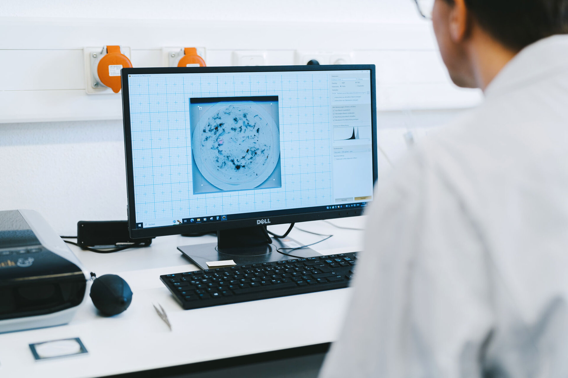 Un employé de laboratoire est assis à son bureau et regarde sur l'écran de son ordinateur une photo d'un module électronique pour l'analyse de la détermination de la pureté. | © @The Sour Cherry Fotografie - Michaela Curtis